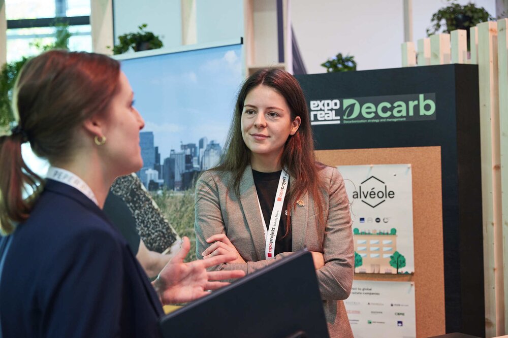 Two women are talking in front of a dark exhibition wall with the logos of EXPO REAL (in white) and Decarb (in green).
