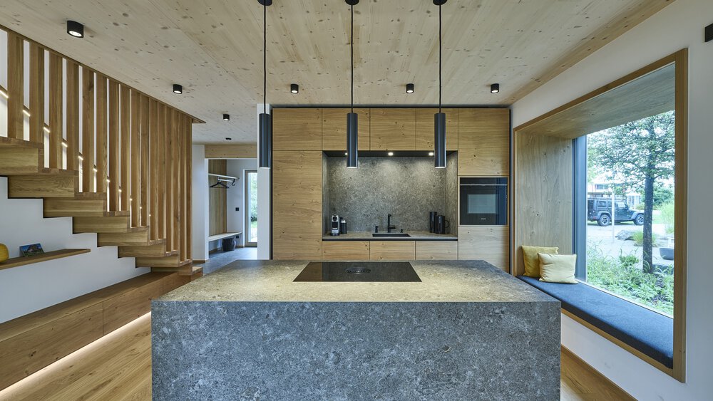 Moderne Küche mit Steininsel, Holzschränken und Hängelampen; großes Fenster mit Sitzgelegenheit auf der rechten Seite.