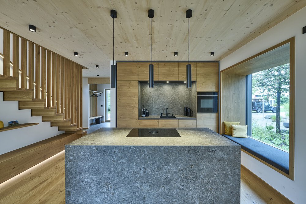 Moderne Küche mit Steininsel, Holzschränken und Hängelampen; großes Fenster mit Sitzgelegenheit auf der rechten Seite.