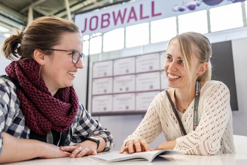Two people sit across from each other at a table discussing an open brochure, with a “Jobwall” displaying job postings at a trade fair in the background.
