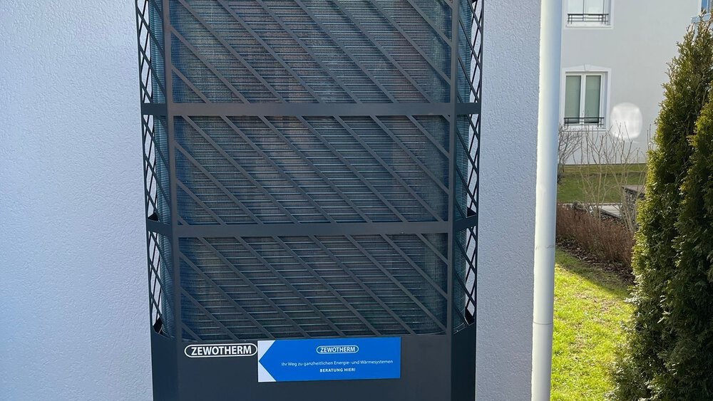 Ein graues Wärmepumpengerät steht auf einem Holzdeck neben einer weißen Wand mit einem Schild darüber.