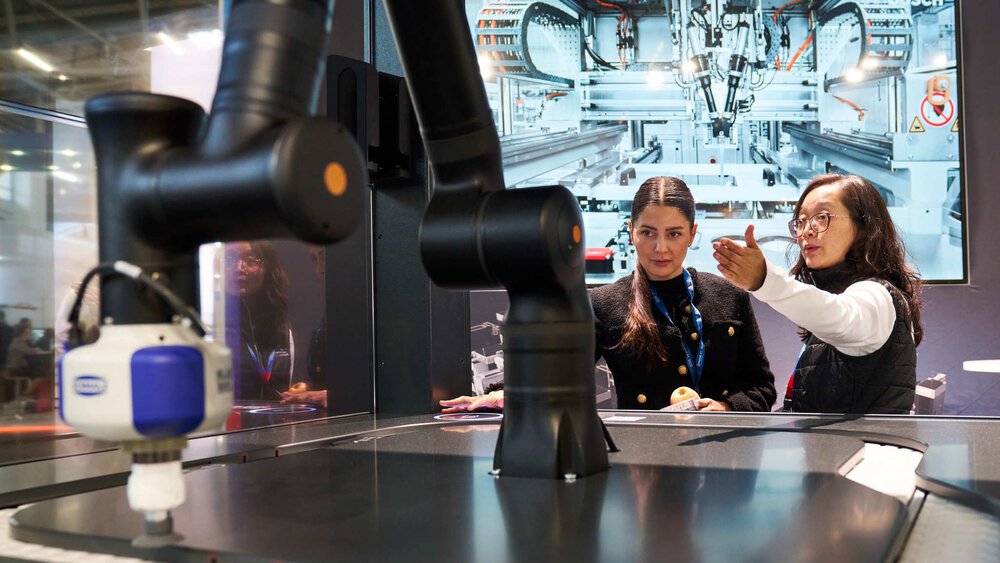 Zwei Frauen stehen an einem Messestand und betrachten einen schwarzen Roboterarm. Auf einem Bildschirm hinter ihnen ist eine weitere Maschine zu sehen.