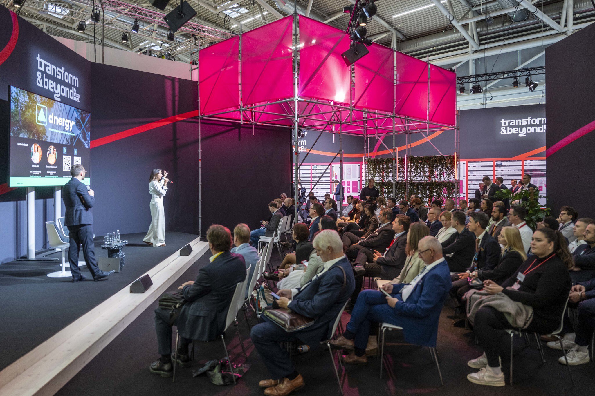 A woman speaks to a seated audience at a technology conference beneath a bright pink geometric structure.