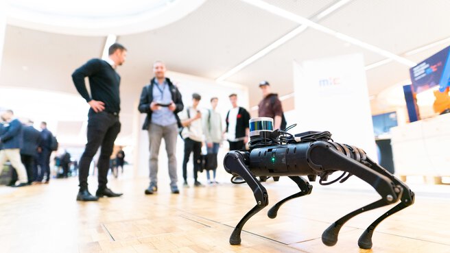 A small four-legged robot stands on a wooden floor while several people look at it from the background.