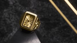 Goldene Uhr mit römischen Ziffern auf schwarzer Marmoroberfläche und diagonalen goldenen Linien im Hintergrund.