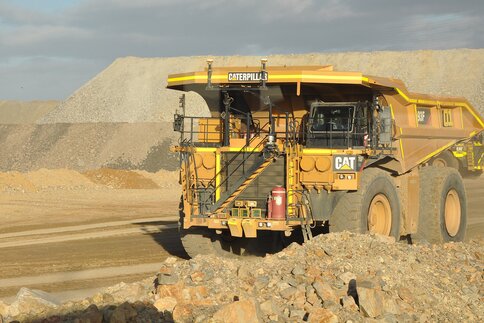 Ein großer gelber CAT-Muldenkipper in einem Kieswerk, mit Schotterhaufen im Hintergrund bei Sonnenschein.