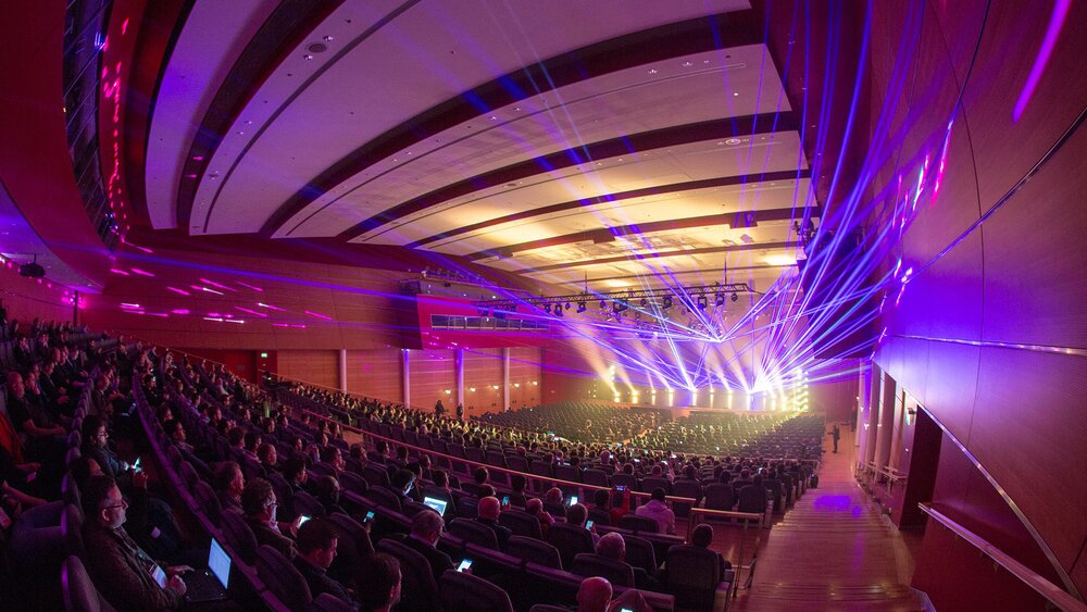 Ein großes Publikum sitzt in einem modernen Saal und blickt auf eine Bühne, über der helle Lichter und bunte Laserstrahlen über die Zuschauer hinwegziehen.