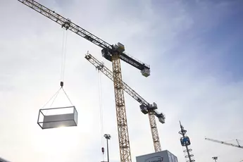 Mehrere große Baukräne heben einen rechteckigen Container in die Luft. Die Sonne scheint durch den Container, und der Himmel ist leicht bewölkt. Im Hintergrund sind weitere Kräne und ein hoher Turm zu sehen.