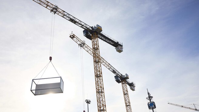 Mehrere große Baukräne heben einen rechteckigen Container in die Luft. Die Sonne scheint durch den Container, und der Himmel ist leicht bewölkt. Im Hintergrund sind weitere Kräne und ein hoher Turm zu sehen.