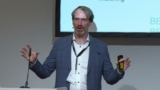 Ein Mann im blauen Anzug hält eine Präsentation und spricht in ein Mikrofon. Im Hintergrund ist ein Bildschirm zu sehen.