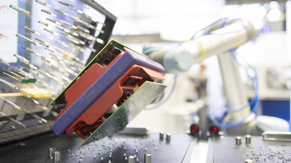 Close-up view of an open machine with circuit boards, metal pins and small cylinders and a white robotic arm in the background.