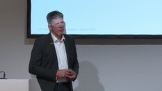 Ein Mann im Anzug hält eine Präsentation. Im Hintergrund ist eine Leinwand mit Text.