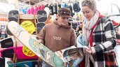 Two people are standing at a trade fair booth with skateboards and balance boards, looking at a brochure together. Several boards and yellow price tags are displayed in the background.