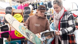 Zwei Personen stehen an einem Messestand mit Skate- und Balanceboards und betrachten gemeinsam einen Prospekt. Im Hintergrund sind mehrere Boards sowie gelbe Preisschilder angebracht.