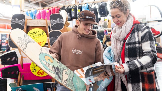 Two people are standing at a trade fair booth with skateboards and balance boards, looking at a brochure together. Several boards and yellow price tags are displayed in the background.