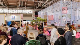 A group of people gather in a brightly lit exhibition hall with colourful stands and decorations.