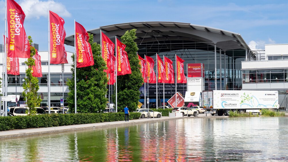 View of the western entrance to the Messe München exhibition centre with the trade fair lake in the foreground and the red flags of transport logistic waving behind it.