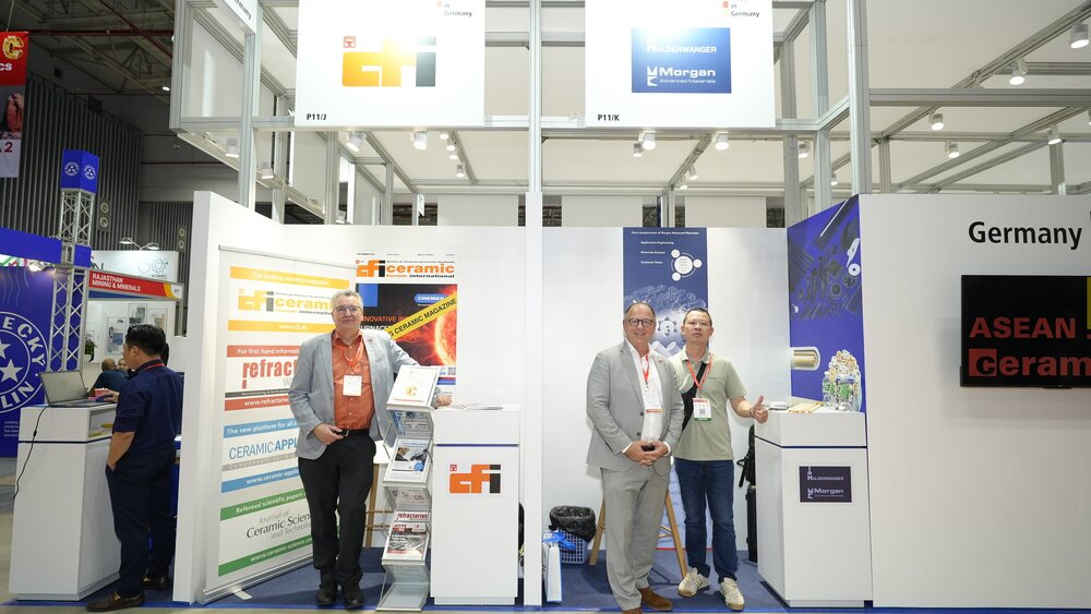 Three men stand at a stand at Asean Ceramics between magazine racks and information displays and pose together for a photo.