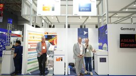 Three men stand at a stand at Asean Ceramics between magazine racks and information displays and pose together for a photo.