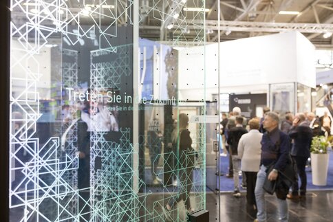 People walk through a trade fair with a glass panel on which the words 