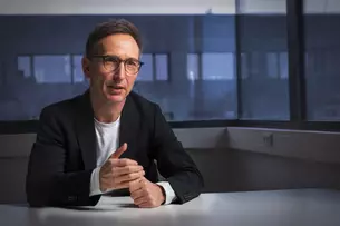 A man with glasses and a suit sits at a table in a dimly lit office with large windows during the interview.