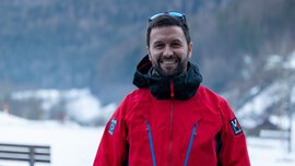 Ein lächelnder Mann mit Bart, kurzen Haaren, Sonnenbrille und einer roten Winterjacke steht in der Natur vor einem bewaldeten Berg im Schnee.