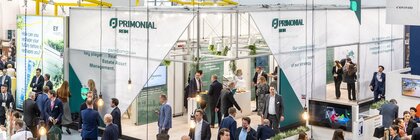 Zahlreiche Besucher schlendern durch eine Messehalle, in der große, moderne Messestände zu aufgebaut sind.