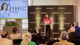 Eine Frau spricht während einer Pressekonferenz auf einem Podium. Auf einem Bildschirm sind ihr Foto und ihr Name zu sehen. Zuschauer machen Fotos.