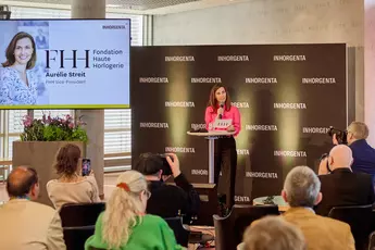 A woman speaks on a podium during a press conference. Her photo and name are shown on a screen. Spectators take photos.