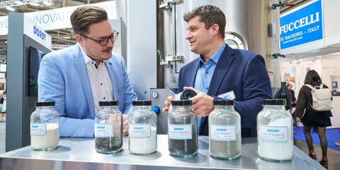 Two trade fair visitors discuss ceramic powder samples at an exhibitor booth.