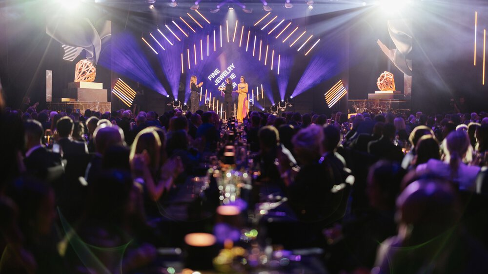 Eine Person spricht auf der Bühne einer glamourösen Preisverleihung vor einem an Tischen sitzenden Publikum.