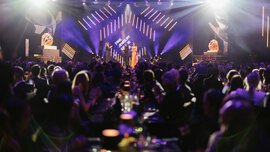 Eine Person spricht auf der Bühne einer glamourösen Preisverleihung vor einem an Tischen sitzenden Publikum.