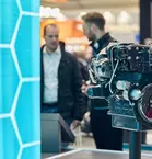 Engine as an exhibit in the exhibition hall in front of a turquoise-blue wall and people in conversation.