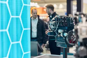 Motor als Ausstellungsstück in Messe-Halle vor türkis-blauer Wand und Menschen im Gespräch.