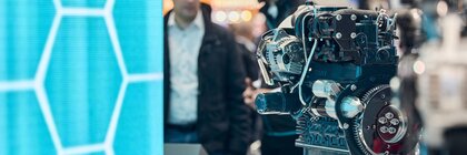 Motor als Ausstellungsstück in Messe-Halle vor türkis-blauer Wand und Menschen im Gespräch.