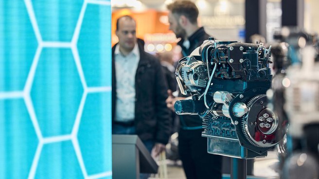 Motor als Ausstellungsstück in Messe-Halle vor türkis-blauer Wand und Menschen im Gespräch.