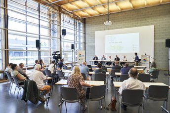 Eine Podiumsdiskussion zu den Poinger Klimatagen in einem hellen Raum mit großen Fenstern vor Publikum und einer Kamera.