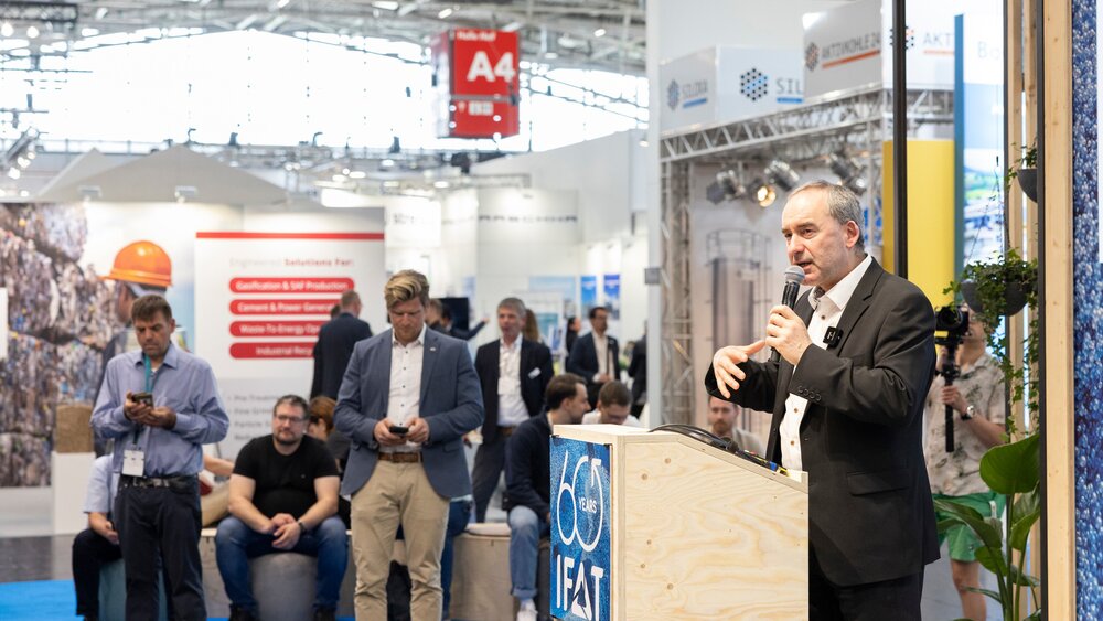 Bavarian Economics Minister Hubert Aiwanger giving a speech at a wooden lectern with the IFAT logo in a busy exhibition hall.