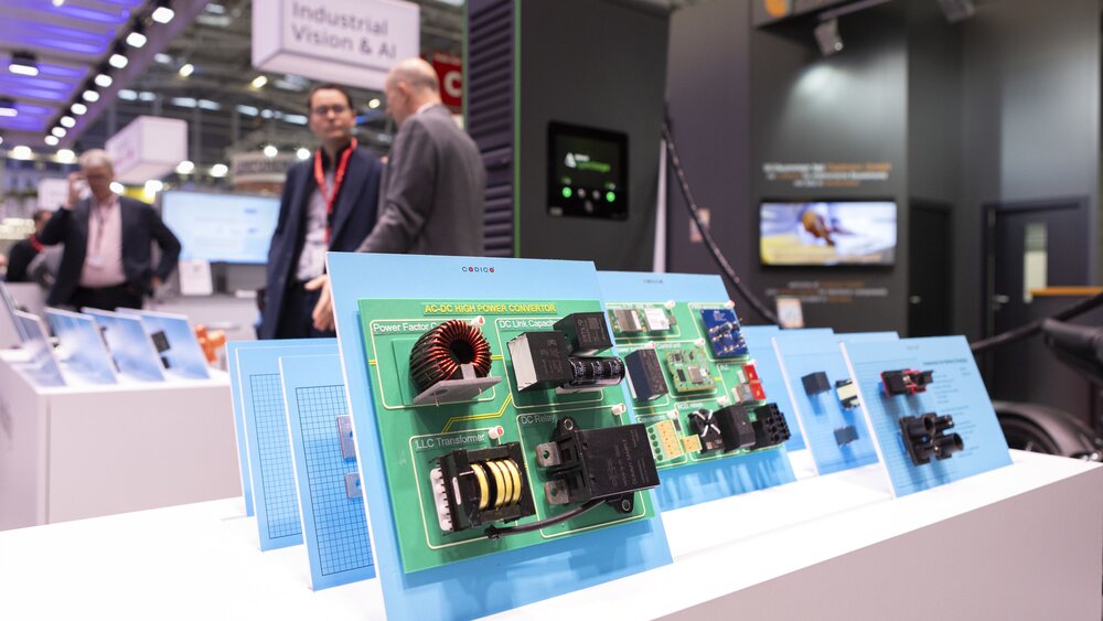 Printed circuit boards and electronic components displayed on pedestals at an exhibition stand. People and exhibition stands can be seen behind them.