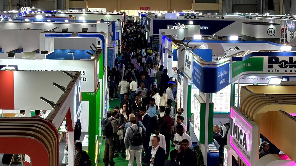 A crowded exhibition hall where people are walking among brightly lit booths and exhibition areas displaying various company signs and products.