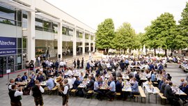 Menschen sitzen an Tischen im Atrium zwischen den Messehallen, umgeben von Bäumen. Im Vordergrund ist eine Blaskapelle zu sehen.