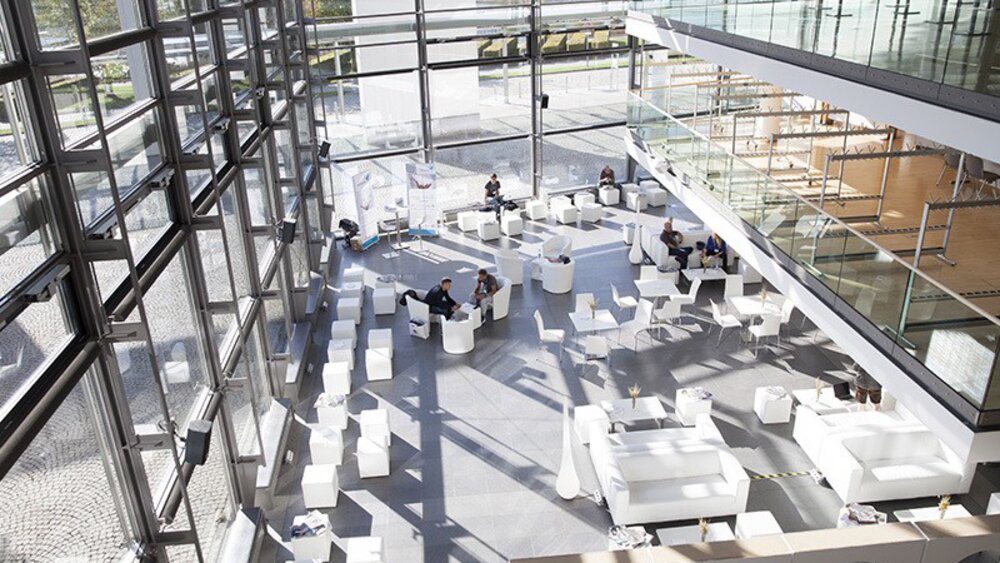 A spacious, modern atrium with large windows, white furniture, and people sitting at scattered tables. Sunlight streams in, casting bright patterns on the floor.