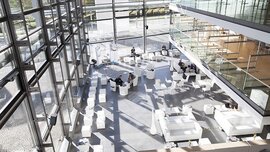A spacious, modern atrium with large windows, white furniture, and people sitting at scattered tables. Sunlight streams in, casting bright patterns on the floor.