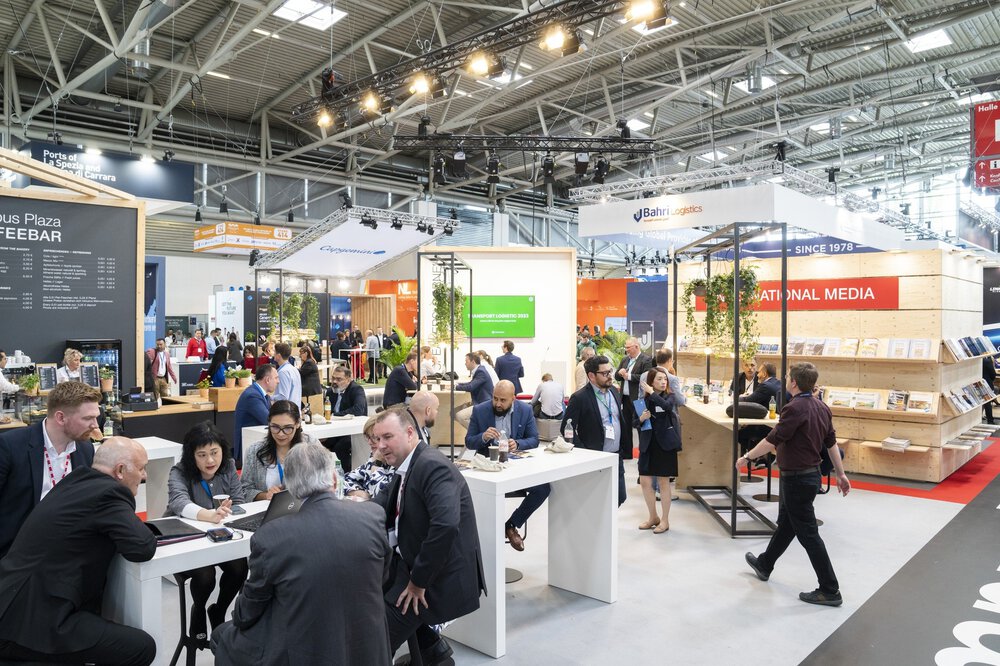 People socialise at a busy trade fair with modern stands and digital displays under industrial ceiling lights.