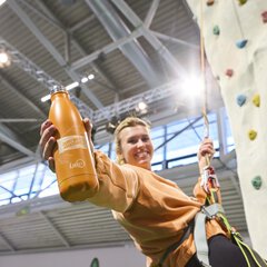 Besucherin erklimmt den Kletterturm der f.re.e 2025 und zeigt ihre Kletterfähigkeiten. Mit ihrer rechten Hand hält sie lächelnd eine orangene Trinkflasche mit f.re.e Branding in die Kamera.
