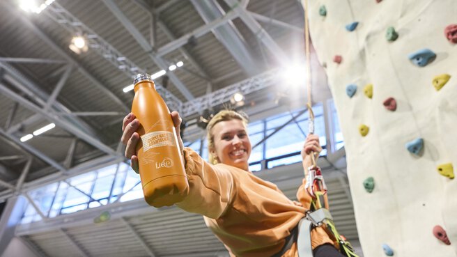 Besucherin erklimmt den Kletterturm der f.re.e 2025 und zeigt ihre Kletterfähigkeiten. Mit ihrer rechten Hand hält sie lächelnd eine orangene Trinkflasche mit f.re.e Branding in die Kamera.