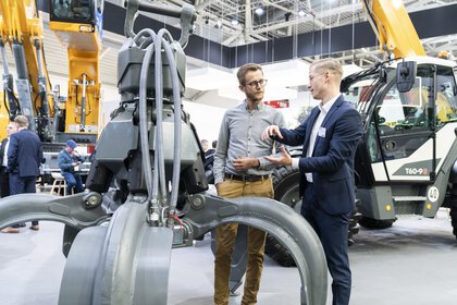 An exhibitor talks to an interested visitor next to the claw of a grab excavator on display.