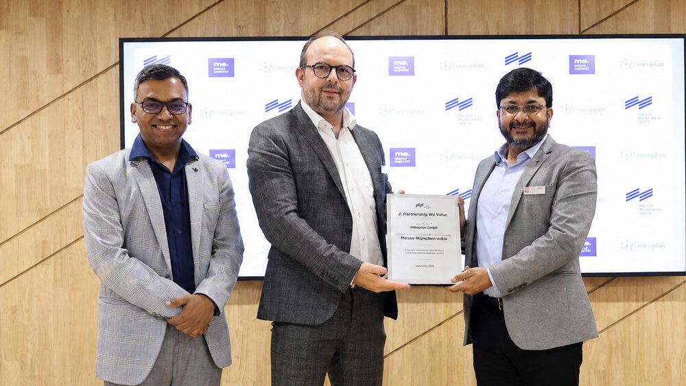 Three men in suits are standing on a stage; two of them are holding a certificate between them and smiling at the camera. Behind them is a presentation board featuring logos.