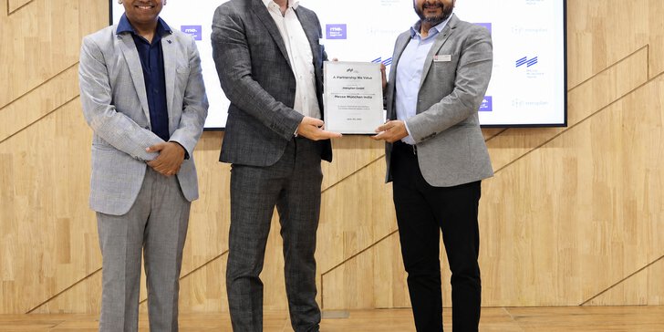 Three men in suits are standing on a stage; two of them are holding a certificate between them and smiling at the camera. Behind them is a presentation board featuring logos.