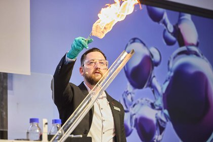 A man in a blazer holds up a metal rod with something burning at the end